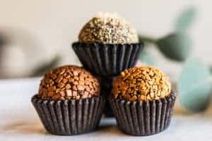 Three assorted chocolate truffles in paper cups are stacked in a pyramid shape. The truffles, crafted by a renowned Chocolate Shop in Durham, have textured coatings in various shades of brown and gold. Two blurred green leaves are in the background, making it perfect for corporate gifts.