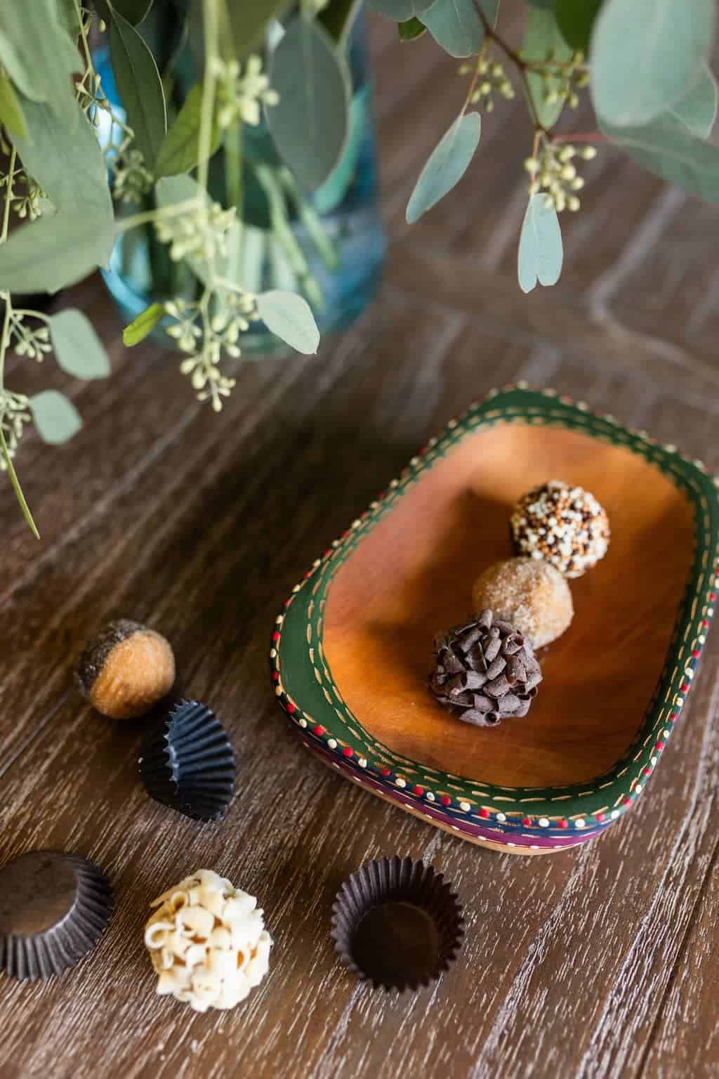 An oblong dish on a wooden surface holds four chocolate truffles, each decorated differently, with three empty wrappers beside it. Greenery is visible in a vase in the background, reminiscent of charming cafes found in places like Raleigh and Durham.