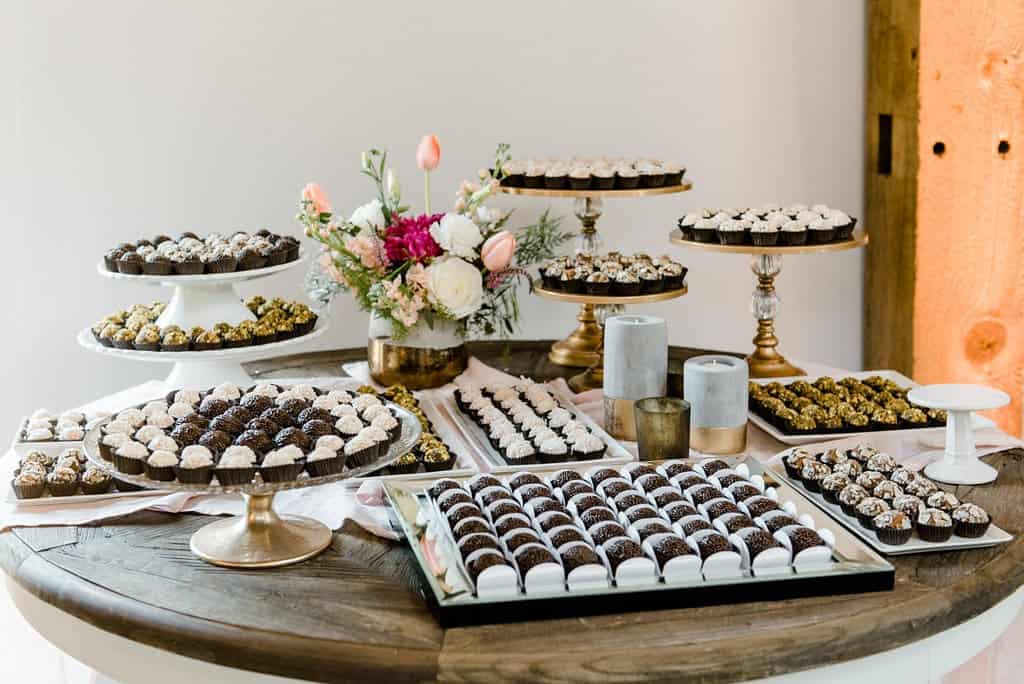 An arranged display of various pastries and treats on a round wooden table, including trays and stands with decorated sweets from a renowned Durham chocolate shop, alongside a floral arrangement.