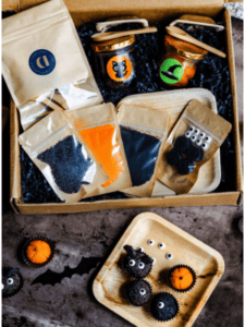 A box containing jars of Halloween-themed craft supplies including containers with googly eyes, black and orange powder, and other small decorations. The items are arranged on and next to a wooden tray.