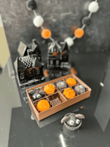 A display of Halloween-themed chocolates, including ones shaped like bats and decorated with googly eyes, set on a reflective surface with black and orange decorations and haunted house figurines in the background.