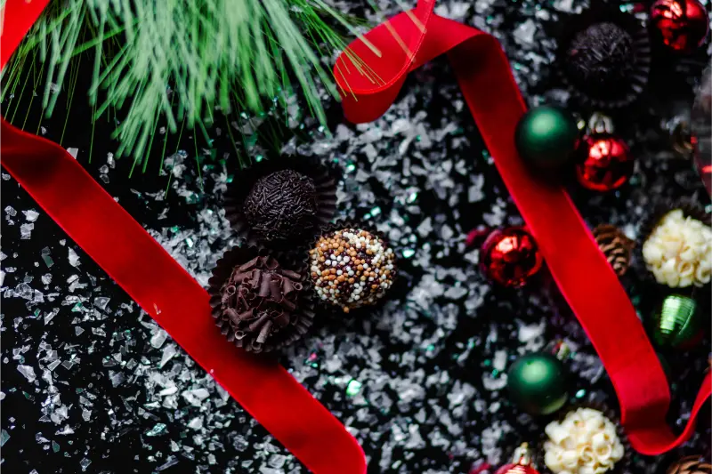 The 8 piece Holiday Gift Box displayed on a snowy surface, adorned with red ribbons, green pine branches, and small red and green ornaments.
