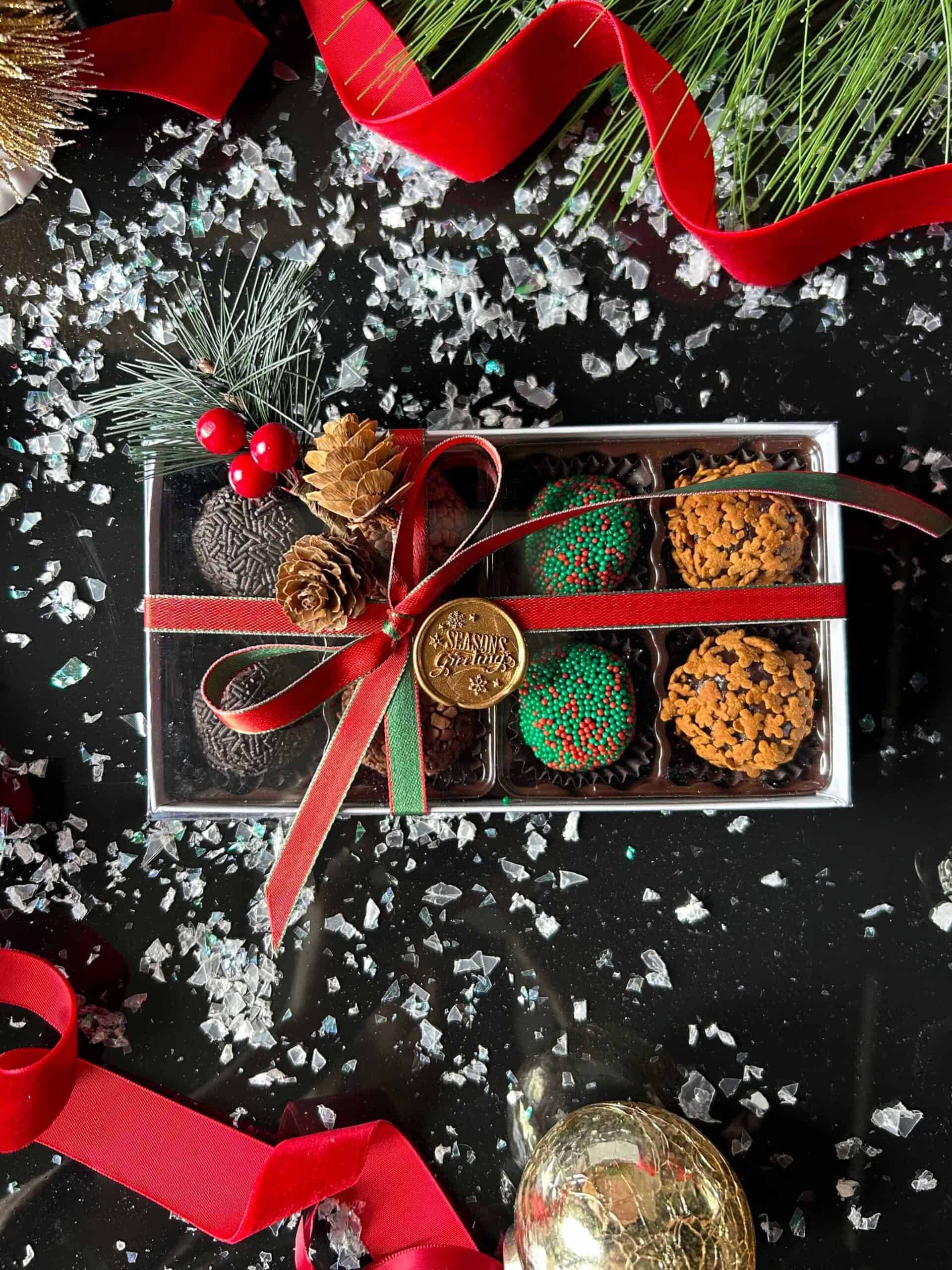 An 8 piece Holiday Gift Box adorned with a red ribbon, nestled among pine cones, red berries, and decorative greenery on a dark surface accented with scattered faux snow and additional red ribbon.