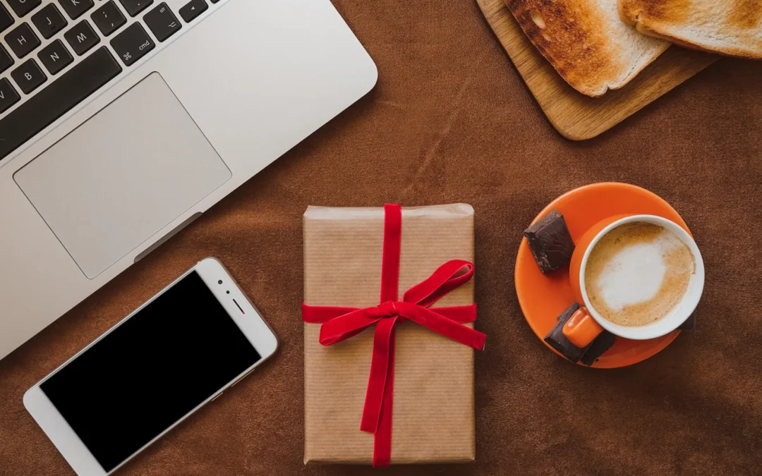 Laptop, smartphone, wrapped gift with a red ribbon, cappuccino in an orange cup, two chocolate brigadeiro pieces, and toast on a wooden board on a brown surface.