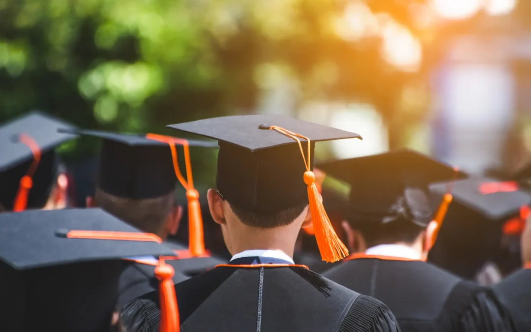 A group of graduates in black caps and gowns with orange tassels stand outdoors, facing away from the camera in sunlight.
